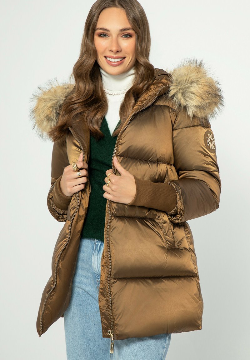 Hnědá puchová bunda s umělou kožešinou na kapuci, prošívaný design, pružné manžety a zip, nošená přes zelený svetr a modré džíny.