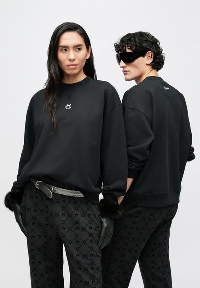 Fekete túlméretezett pulóverek kerek logó részlettel, fekete mintás nadrággal, mely finom mintázattal rendelkezik. Fekete kesztyűk és öv díszítések is tartoznak hozzá.