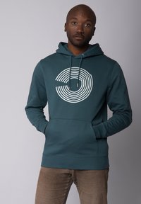 Tegenvloeiende hoodie in teal met een witte geconcentreerde cirkelsgrafiek, verstelbare trekkoordkap en een voorzak; zachte textuur en aansluitende manchetten.
