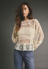 Blusa trasparente color crema con motivi floreali testurizzati e dettagli in pizzo, caratterizzata da maniche a palloncino e orlo arricciato. Indossata con jeans blu.