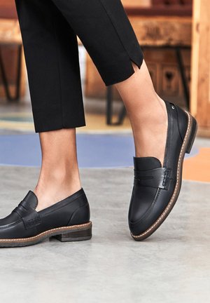 Mocassins en cuir noir avec un talon bas, présentant des coutures décoratives et une finition lisse. Portés avec un pantalon noir court, mettant en valeur la cheville.
