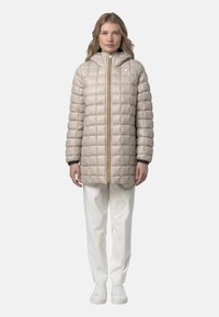 Cappotto imbottito color beige con cerniera frontale, cappuccio e polsini a coste. Presenta un design trapuntato e una cerniera dorata. Indossato sopra pantaloni bianchi e sneaker.