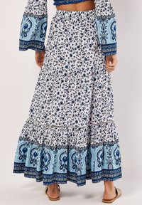 Jupe maxi florale blanche, ornée de motifs floraux bleus, avec un ourlet volanté en niveaux et une taille élastique. Portée avec des sandales.
