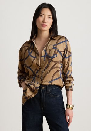 Frau mit langen schwarzen Haaren, die ein hellbraunes Hemd mit blauen und braunen Gürtelmustern, dunkle Jeans und ein goldenes Manschettenarmband trägt, steht mit einer Hand in der Tasche.