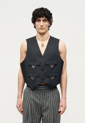 Young man with curly hair wearing a black sleeveless vest with four flap pockets and striped pants, standing against a plain background.