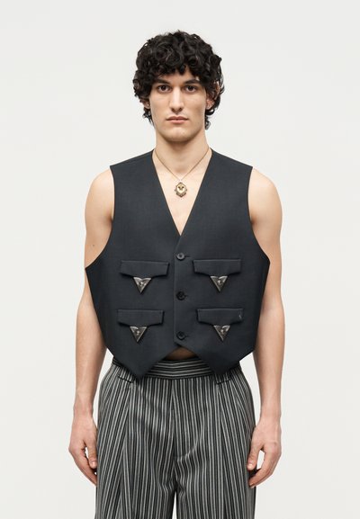 Jeune homme aux cheveux bouclés portant un gilet noir sans manches avec quatre poches à rabat et un pantalon rayé, debout devant un fond uni.