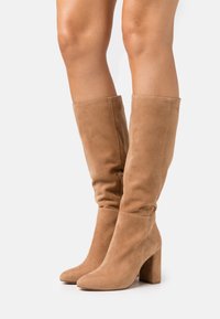 Bottes hautes jusqu'au genou en daim couleur noisette, avec un bout arrondi et un talon bloc. Les bottes présentent un design décontracté pour un ajustement ample autour du mollet.