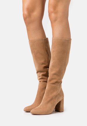 Bottes hautes jusqu'au genou en daim couleur noisette, avec un bout arrondi et un talon bloc. Les bottes présentent un design décontracté pour un ajustement ample autour du mollet.