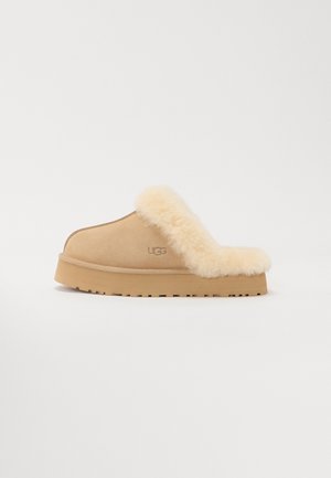 Beige suede slipper with a fluffy shearling collar and a light sole. Features a rounded shape and the UGG logo embossed on the side.