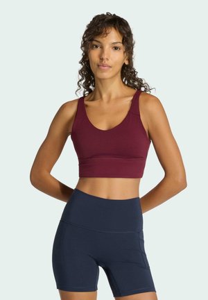 Vínová sportovní podprsenka se širokými ramínky a kulatým výstřihem, kombinovaná s tmavě modrými kraťasy s vysokým pasem, které mají hladký povrch a minimální švy.