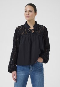 Sort bluse med et blondernederdel og en flæsekrave, sløjfe-lukning og lange ærmer med elastiske manchetter. Båret med blå jeans.