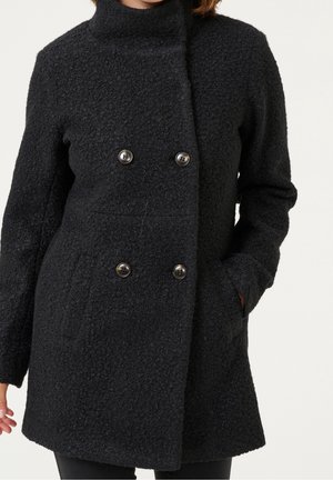 Femme portant un manteau en laine noire texturée à double boutonnage, une main dans la poche, sur un fond neutre.