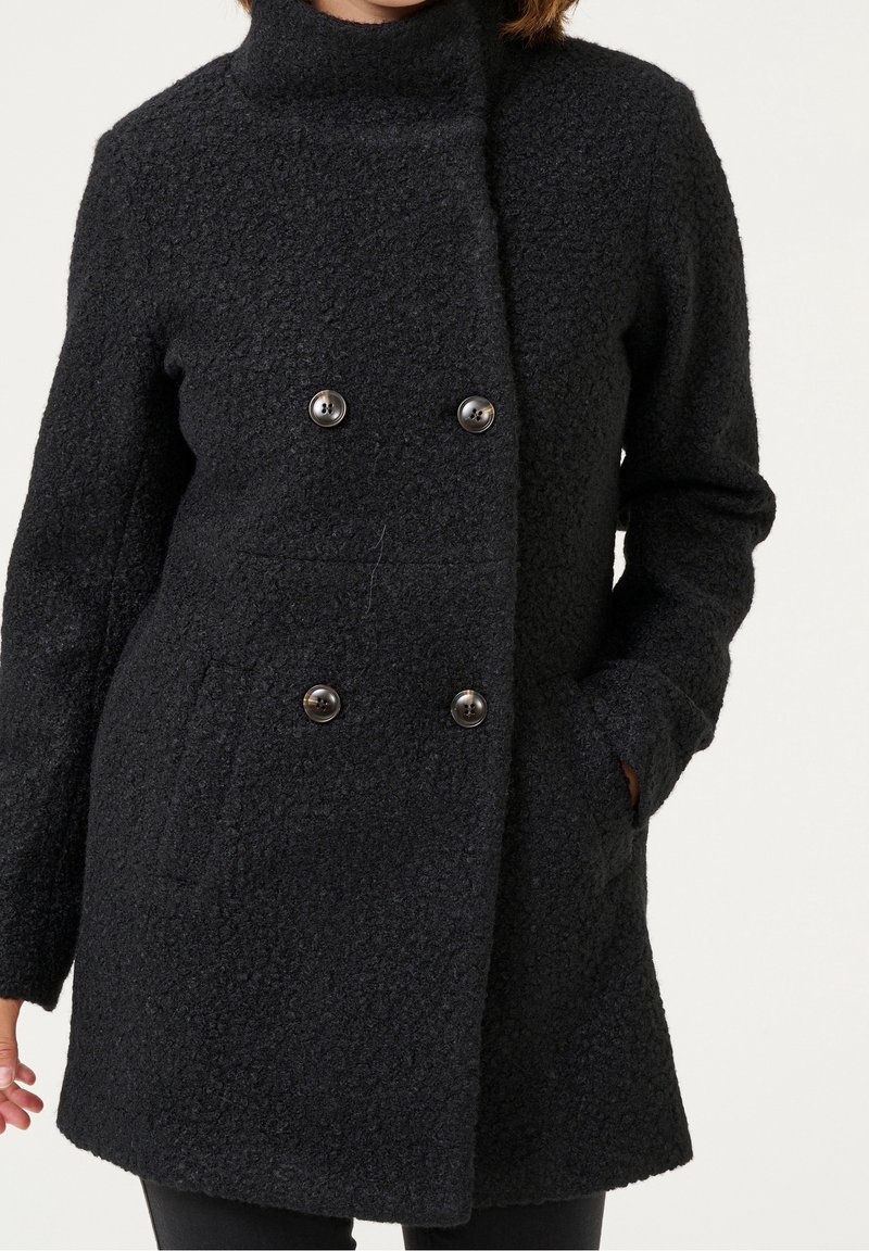 Femme portant un manteau en laine noire texturée à double boutonnage, une main dans la poche, sur un fond neutre.