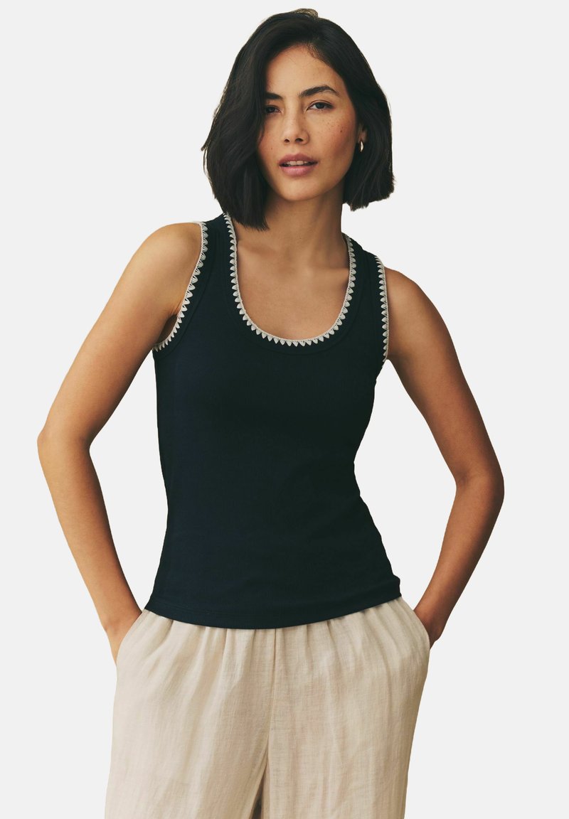 Sort tanktop med ribbet tekstur, udsmykket halsudskæring med hvid detaljering, parret med lyse linnedbukser.
