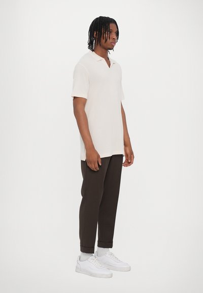 Polo shirt beige clair à manches courtes, col en V et coupe décontractée, associé à un pantalon tapered marron foncé et des baskets blanches.