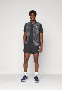 Mann in schwarzem Sportweste, schwarzem T-Shirt, schwarzen Shorts, weißen Socken und schwarzen Laufschuhen vor einem schlichten Hintergrund stehend.