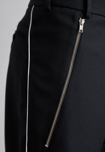 Pantalons en tissu noir avec une fermeture zippée sur le côté dotée de dents en métal doré et une fine bande blanche le long de la couture.