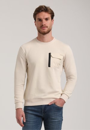 Light beige sweatshirt with long sleeves, ribbed collar and cuffs, featuring a black zippered chest pocket. Paired with blue jeans.