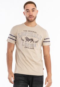 Beige bomulds t-shirt med kortærmer. Har et trykt logo med en løve, tekst og to brune striber på ærmerne.