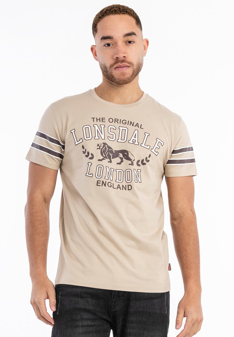 Beige bomulds t-shirt med kortærmer. Har et trykt logo med en løve, tekst og to brune striber på ærmerne.