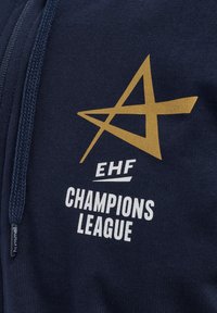 Marinblå hoodie med en guldstjärnlogotyp, med "EHF" och "CHAMPIONS LEAGUE" i vit text. Mjukt texturerat material med zip-detail.
