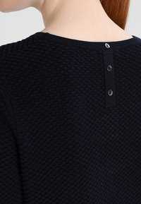 Marinblå texturerad stickad tröja med rund halsringning och tre knappstängningar bak med svarta knappar.