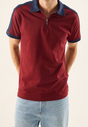 Homme portant un polo rouge à manches courtes avec col et bandes aux épaules bleu marine, associé à un jean bleu clair, les mains dans le dos.