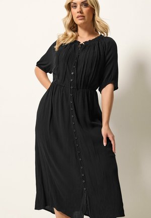 Blonde vrouw draagt een zwarte midi-jurk met knopen, korte mouwen en een gestructureerde stof, poserend tegen een lichte achtergrond.