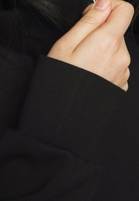 Svart långärmad topp med hög krage, med en slät yta och åtsittande design. Närbilden visar manschettdetaljen och tygkvaliteten.