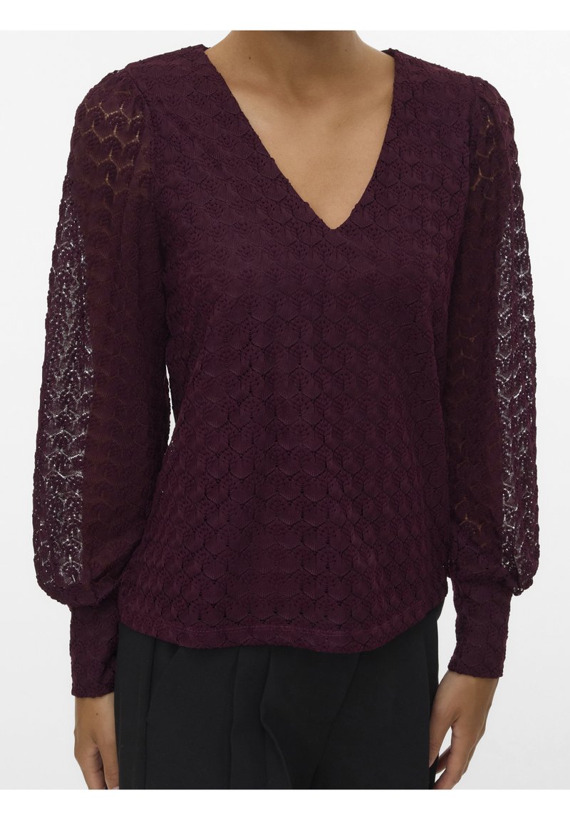 Femme portant un chemisier en dentelle violet foncé avec de longues manches bouffantes transparentes et un décolleté en V, associé à un pantalon noir.