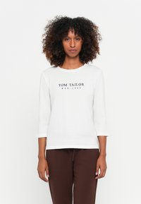 Witte shirt met driekwart mouwen met "TOM TAILOR EST. 1962" in donkere letters gedrukt op de voorkant, gecombineerd met bruine broeken.