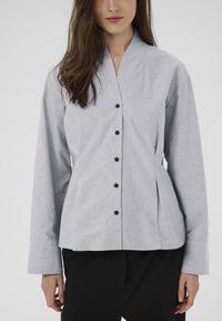 Kvinde med langt brunt hår iført en lysegrå knappet jakke med V-udskæring og sorte bukser, stående foran en ensfarvet baggrund.