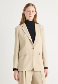 Beige tailored blazer with a single button, notched lapels, and two front pockets, paired with a black turtleneck. Smooth texture.