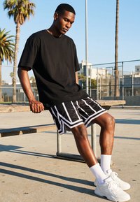 Tânăr în tricou negru și pantaloni scurți cu model, așezat pe o bancă de lemn, pe un teren de baschet în aer liber, sub cer senin.