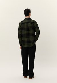 Vyriškis stovintis nugara į priekį, dėvintis žalią ir juodą languotą marškinius, juodus kelnes ir rudos spalvos batus, prieš paprastą šviesią foną.