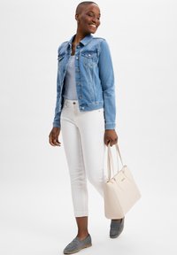 Lichtblauwe denimjack over een grijze top, witte slim-fit jeans, grijze instapschoenen en een beige handtas met gouden accenten.