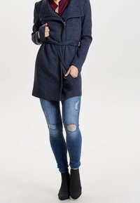 Cappotto in misto lana blu navy con chiusura a zip, ampio colletto e vita con cintura; abbinato a jeans skinny blu con effetto usurato e stivaletti neri alla caviglia.