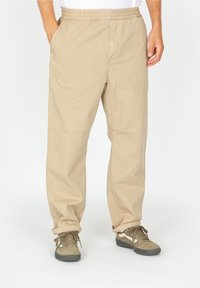 Beige, elastic-waist pants with a loose fit, featuring two side pockets and a smooth cotton texture, paired with olive-green shoes.