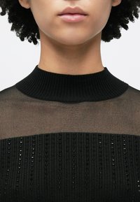 Μαύρο πουλόβερ με ψηλό ριμπ λαιμό και διαφανή πάνω περιοχή. Διαθέτει υφασματινές οριζόντιες ρίγες με μικρές χάντρες κατά μήκος του κάτω μέρους.