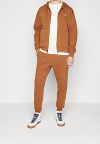 Felpa marrone e pantaloni da jogging realizzati in un tessuto morbido. Design con cappuccio e tasche frontali, di colore coordinato. T-shirt bianca indossata sotto.