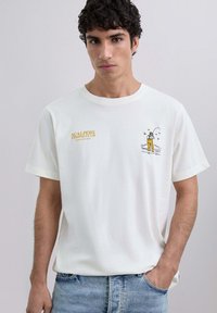 Tânăr îmbrăcat cu un tricou alb cu text galben "Scalpers Fishing Club" și o ilustrație de pescuit, asortat cu blugi albaștri deschis.