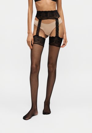 Femme portant des sous-vêtements beige avec jarretelle en dentelle noire et bas résille noirs debout devant un fond uni.
