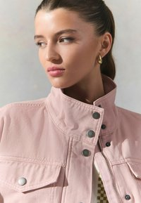 Veste en denim rose pâle avec un col haut, boutons-pression en métal argenté, et deux poches avant ; présente un tissu texturé et une finition lisse.