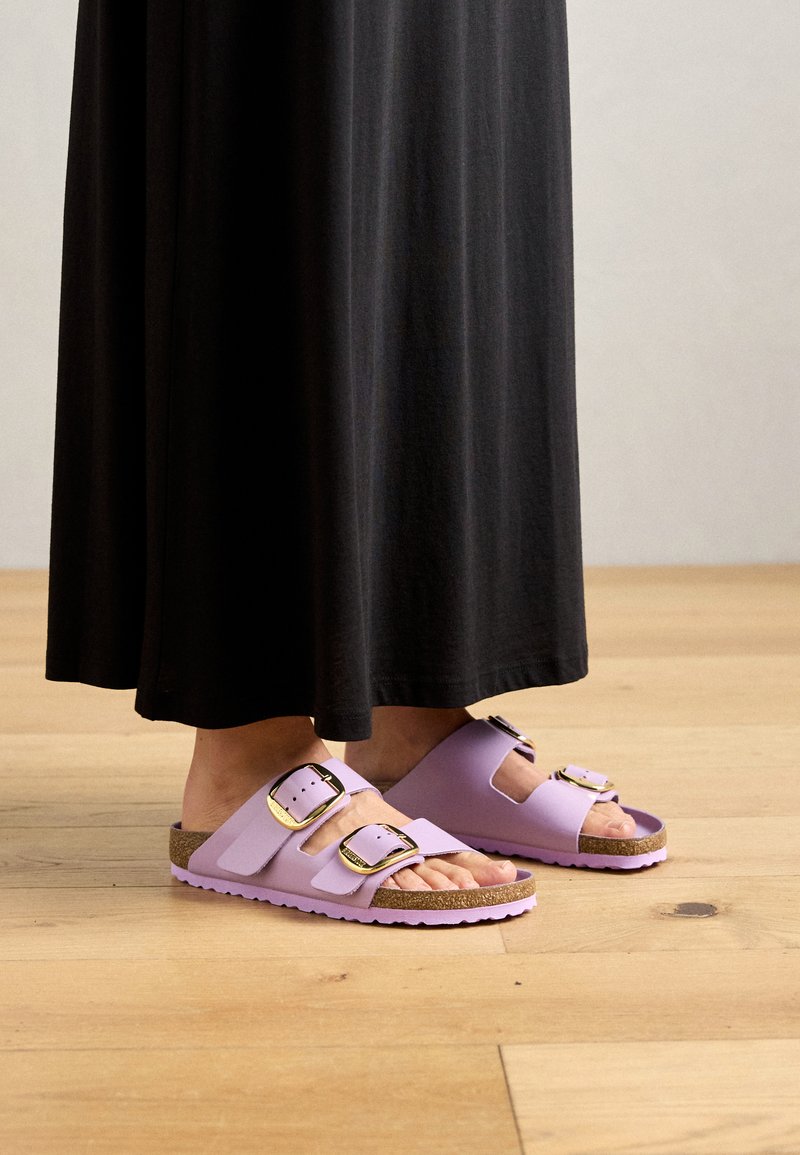 Person in einem schwarzen langen Rock und helllila Sandalen mit zwei großen goldenen Schnallen, die auf einem hellen Holzboden stehen.