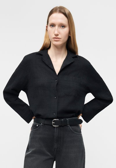 Kvinne med langt, rett hår iført en svart skjorte med knapper, brettet inn i mørkegrå jeans med svart belte, stående med hendene på hoftene.