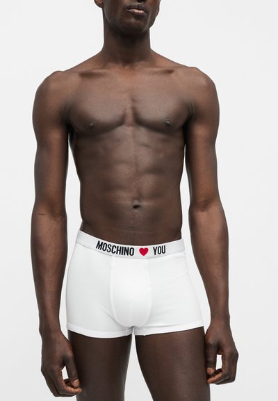 Mandlig model med mørk hud iført hvide Moschino boxershorts med logo og rødt hjerte på linningen, stående mod en ensfarvet lys baggrund.