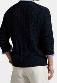 Pull en maille torsadée bleu marine avec ourlet et poignets côtelés. Porté sur une chemise rayée bleu clair. Design classique et texturé.