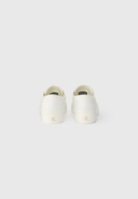 White canvas sneakers with a low-top design and textured fabric. Branded heel accent visible. Minimalist style with a clean look.