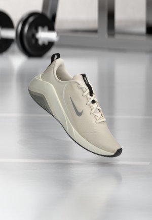 Λευκό αθλητικό παπούτσι Nike με γκρι swoosh, μαύρες λαβές και υφασμένο πάτο, αιωρούμενο πάνω από γυαλιστερό δάπεδο γυμναστηρίου με θολά βάρη στο φόντο.