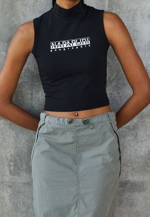 Person trägt ein schwarzes ärmelloses Crop-Top mit dem Text "NAPAPIJRI geographic" und einen hoch taillierten hellgrauen Rock vor einer Betonwand.